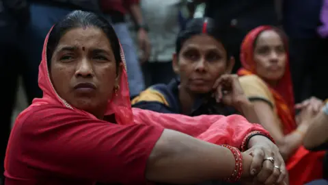 Reuters Three women factory worker - wearing saree - sit, as they participate in a protest demanding wage hikes from their company, in Haryana's Manesar on 7 April 2026. 