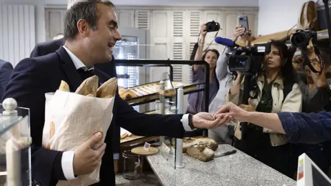 AFP via Getty Images French Prime Minister Sébastien Lecornu (left) pays for baguettes during a visit to a village bakery in Saint-Julien-Chapteuil, central France. Reporters (on the right) are seen filming the prime minister. Photo: 1 May 2026