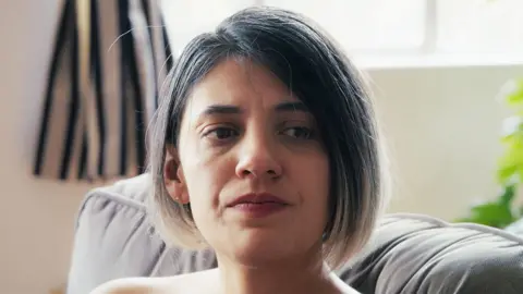 Alex has balayage hair cut in a bob and has dark eyes and wears red lipstick - she is sitting on a sofa and looks mournful