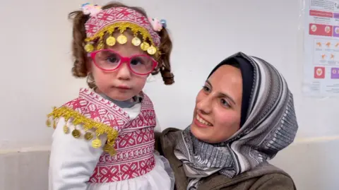 BBC Mother stands (on right) looking up smiling towards her toddler daughter who she is holding. The mother is wearing a headscarf and the daughter red-and-white traditional embroidered dress and pink glasses.