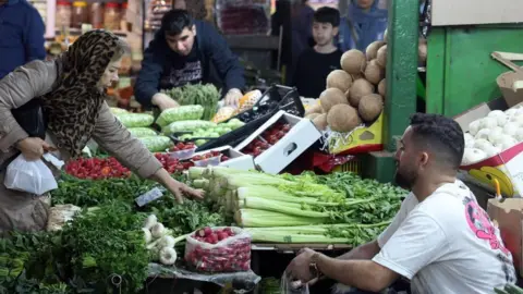 EPA A woman leans across a vegetable stall as a seated man watches her, at a market in Tehran (24/03/26)