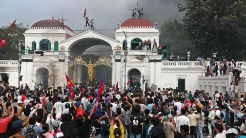 Getty Images Hundreds of protesters gather at the Singha Durbar, the main administrative building for the Nepal government. Some are seen waving the national flag and some are standing on the roofs of the building. A cloud of smoke is billowing in the background