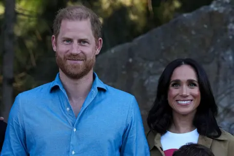Getty Images Harry stands next to Meghan. He is wearing a light blue shirt. They are both smiling.