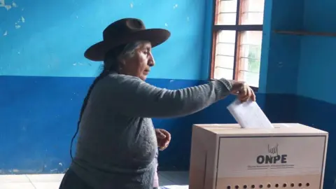 Sengo Perez/EPA/Shutterstock A woman with her hair in plaits and wearing a hat and a woolly jumper casts her vote during the presidential election in Urumbamba, Cusco, Peru, 12 April 2026. She is holding the ballot in her hand and is inserting it into a ballot box. 