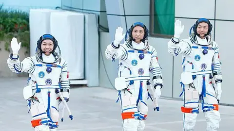 Getty Images Three Chinese astronauts wearing white space suits wave in front of a jubilant crowd waving Chinese flags ahead of the Shenzhou-23 launch