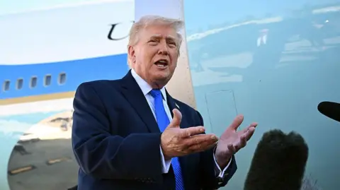 Getty Images Trump wearing a suit and blue tie gesturing with his hands in front of microphones