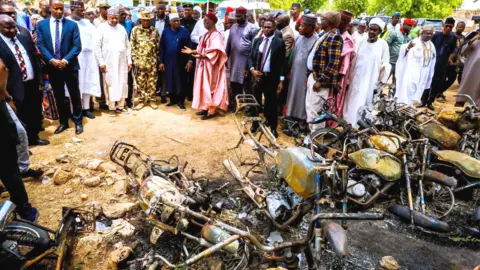 Adamawa State Government A group of men and officials looking on at the burnt-out remnants of motorbikes 