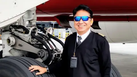 Capt Sumeet Sabharwal's family Capt Sumeet Sabharwal stands next to a wheel of a plane wearing a dark coloured pullover over his pilot's uniform and reflective glasses.
