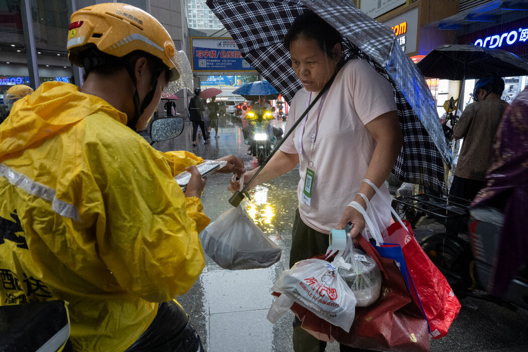 在賽格廣場外，一名騎電動車的外賣騎手將餐食交給一位代送跑腿員，由他送到大樓裡下單的顧客手中。