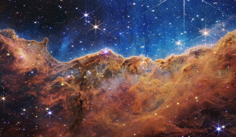 A vibrant image of the Carina Nebula shows bright stars scattered across a deep blue space background, with swirling clouds of orange and brown interstellar dust forming a jagged, mountainous ridge.