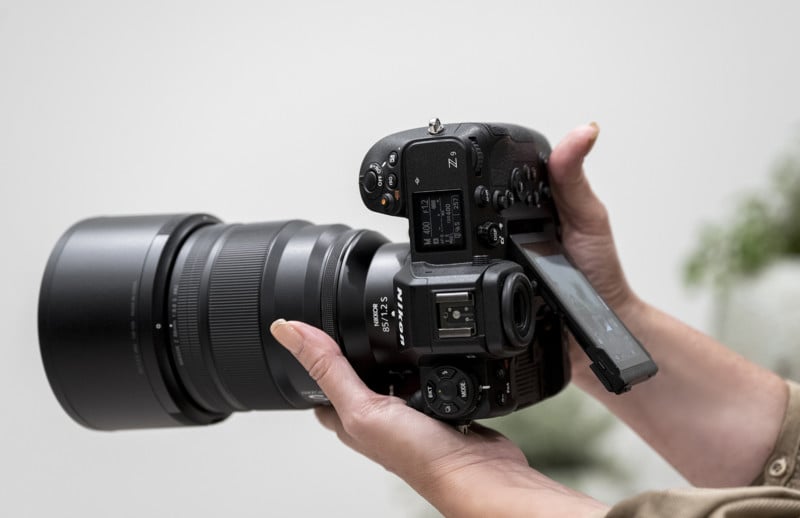 A person holds a professional digital camera with a large lens and a flip-out screen, preparing to take a photo. The camera buttons and dials are visible, as well as the person's hands and part of their sleeve.