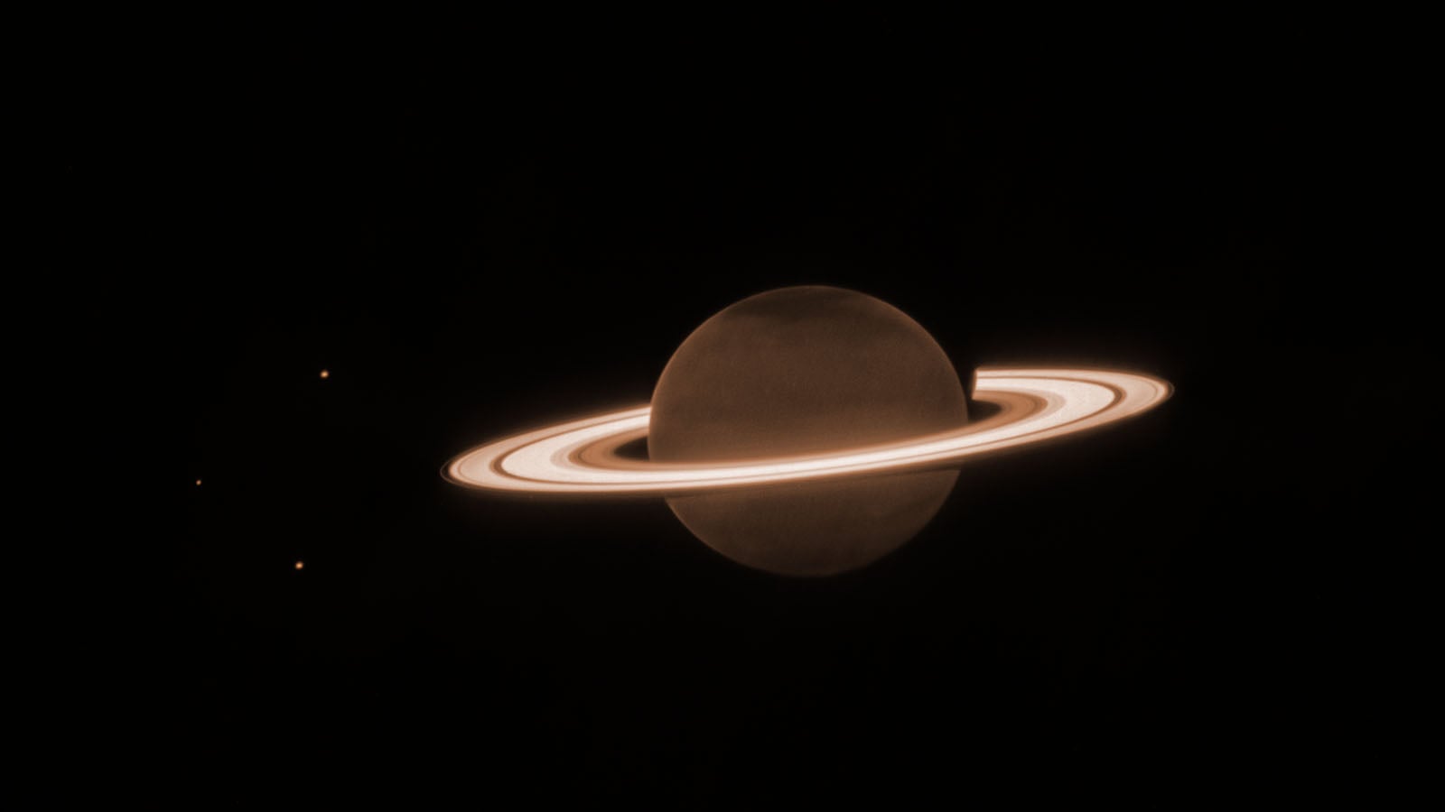 Saturn with its bright rings is seen against a dark background. Three small, faint dots, likely moons, appear to the left of the planet. The overall color tone is a muted brownish-orange.