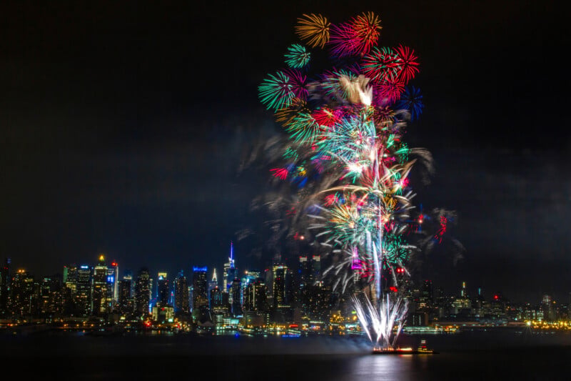 Fireworks in vibrant colors explode over a city skyline at night, illuminating the buildings below with bright, colorful bursts against the dark sky.