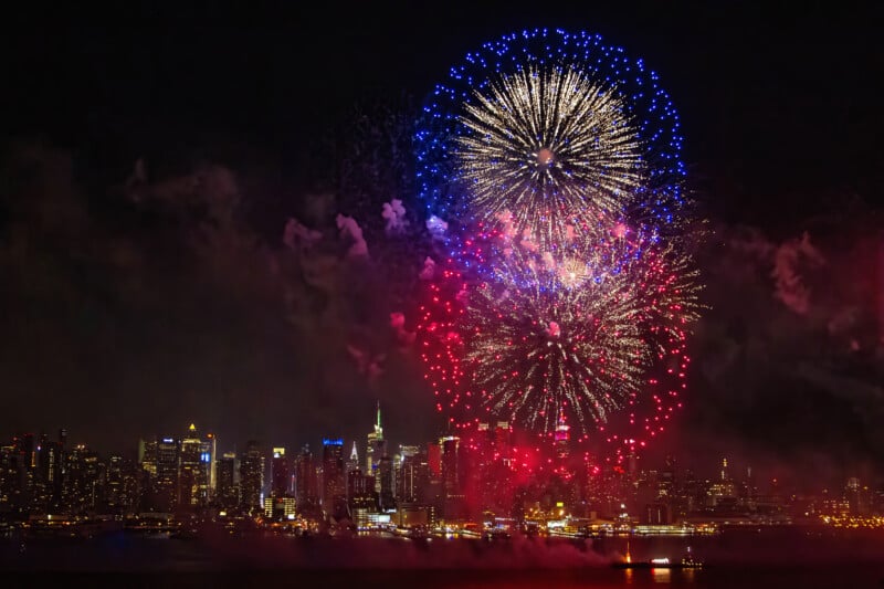 Fireworks in red, white, and blue explode over a city skyline at night, illuminating buildings and reflecting on the water below.