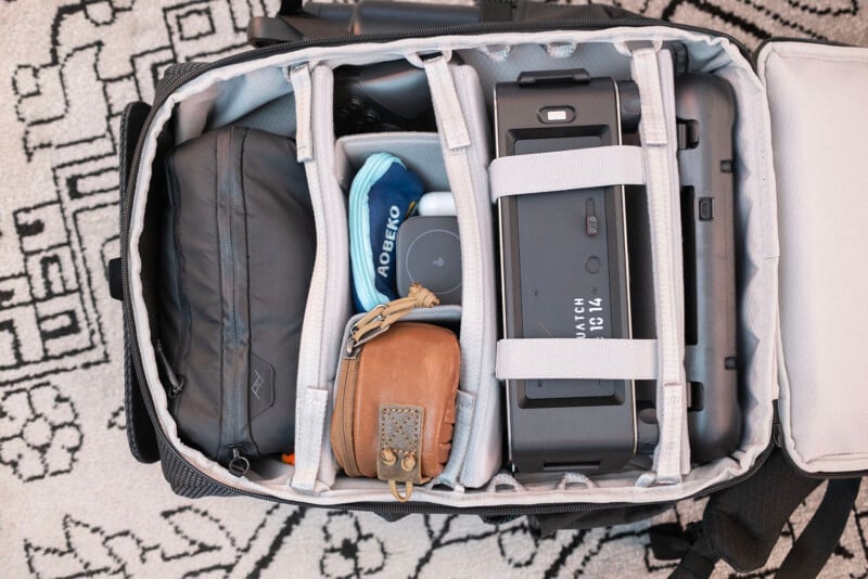 An organized camera backpack containing pouches, a camera case, a brown leather wallet, a blue cloth, and various accessories, all neatly separated by padded dividers. The bag is open on a patterned rug.