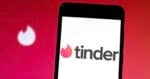 A smartphone displays the Tinder app logo on its screen, with a blurred Tinder flame icon in the background against a red gradient backdrop.