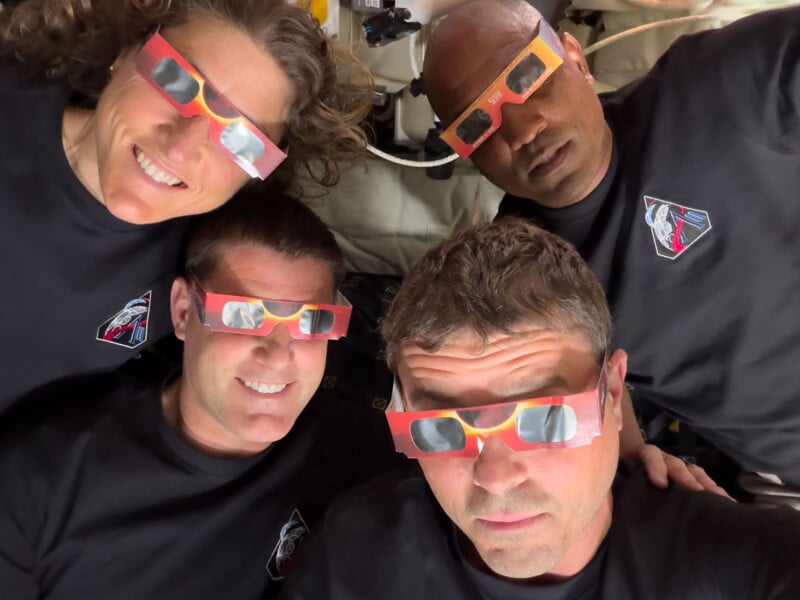 Four people wearing matching black shirts and colorful eclipse glasses smile and look up at the camera, appearing to be in a spacecraft or space station setting.