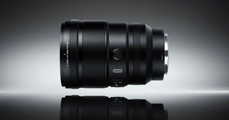 A black camera lens is positioned horizontally on a reflective surface against a gradient gray background, displaying its details and metallic lens mount.