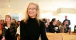 A woman with long blonde hair and glasses, wearing a black turtleneck, sits smiling in a brightly lit room with people seated in the background.