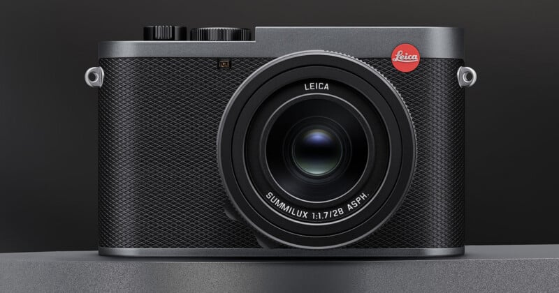 A close-up view of a black Leica digital camera with a textured grip, prominent lens, and the red Leica logo on the top right corner, set against a dark background.