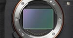 Close-up view of a digital camera's exposed sensor and metal lens mount, showing electronic contacts and screws, with part of the camera body visible on the left and a number 
