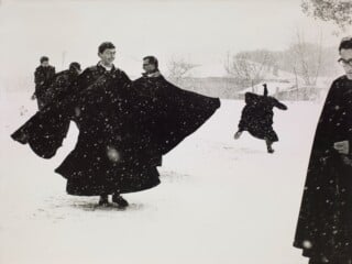 Several men in long black cassocks play in the snow, with one figure leaping in the background and another standing in the foreground.
