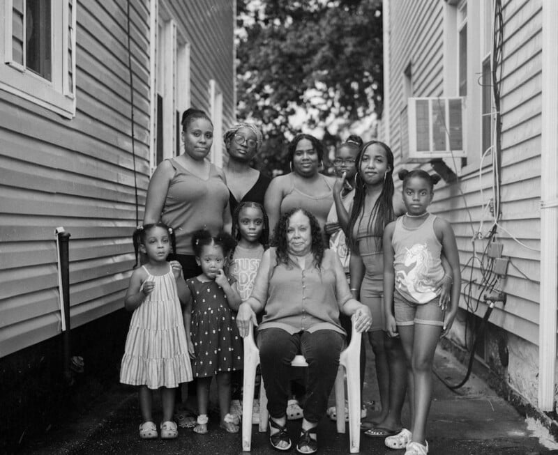 A group of women and young girls pose together between two houses. One older woman sits in a white chair at the center, surrounded by others standing, all looking at the camera.