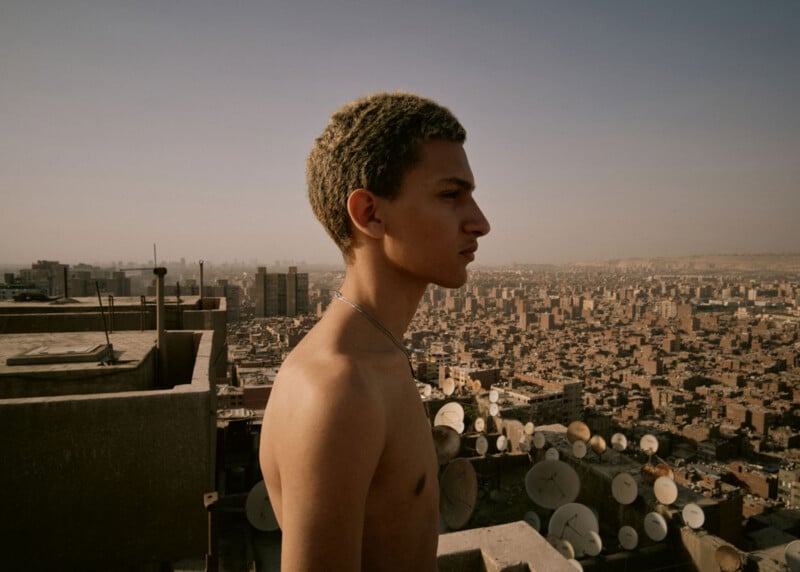 A shirtless young man with short, curly hair stands on a rooftop, looking out over a sprawling city filled with buildings and satellite dishes under a hazy sky.