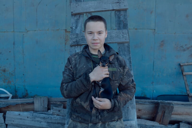 A person in a camouflage jacket stands in front of a wooden ladder and blue wall, holding a small black and tan dog.
