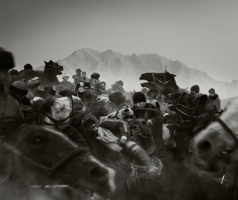 A group of people on horseback compete intensely in a traditional Central Asian game; dust swirls around them, and mountains rise in the background.