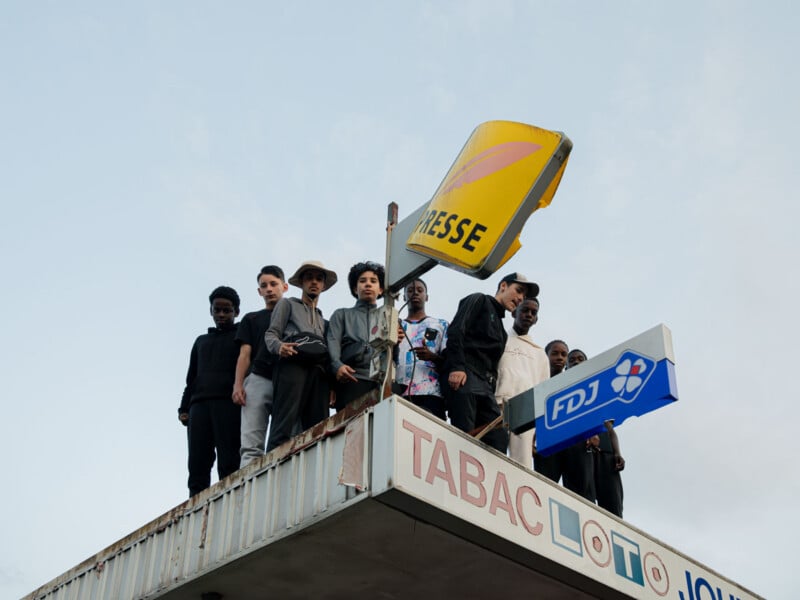 A group of people stand on the roof of a building beneath a yellow "PRESSE" sign. The building has "TABAC" and "FDJ" signs visible. The sky is clear in the background.