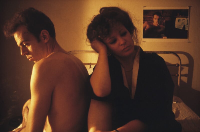 A man and a woman sit back-to-back on a bed in a dimly lit room, with a framed photograph of a man hanging on the wall behind them.