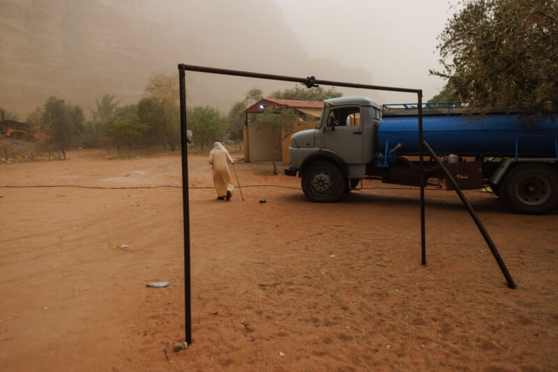 A person in a light robe walks away from a blue water truck on sandy ground, with trees and small buildings in the background under a hazy sky.