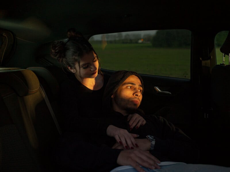A woman sits in a car's backseat, gently holding a man lying across her lap. Both look calm and relaxed, with warm sunlight softly illuminating their faces.