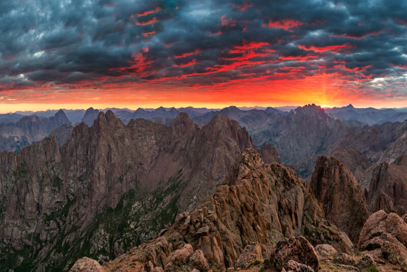 Rocky mountains under a dramatic sunset sky with dark clouds and vivid red-orange light at the horizon, highlighting rugged peaks and deep valleys.