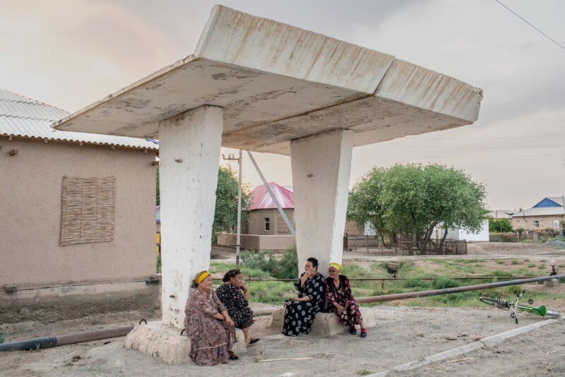 Four women sit and talk under a large concrete structure with a flat roof, near houses and trees, on a quiet, rural street. A bicycle lies on the ground nearby.