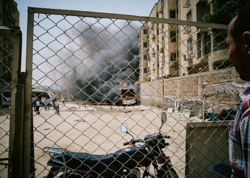 A large plume of black smoke rises from a burning vehicle in a fenced lot between buildings, with a motorcycle in the foreground and a person observing the scene.