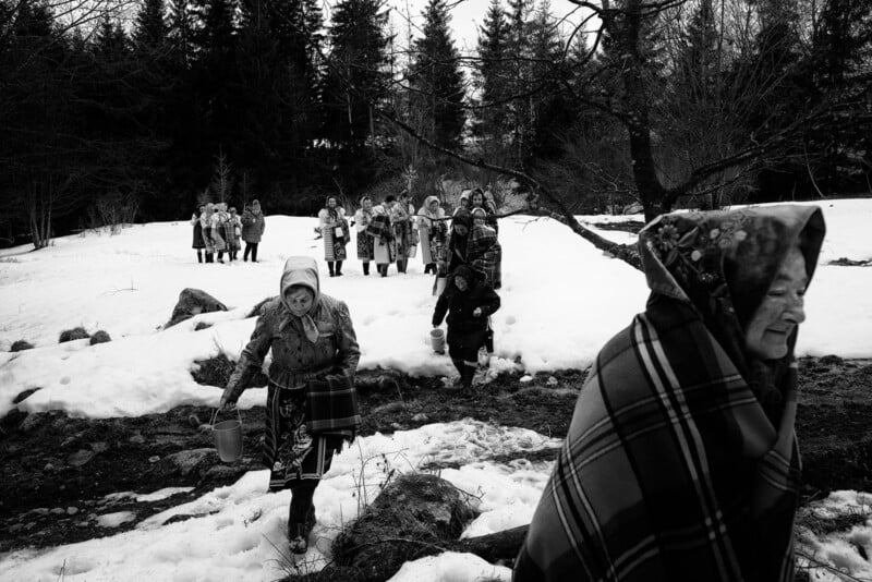 A group of women in headscarves walk through a snowy landscape, some carrying buckets and wearing patterned clothing, with trees and rocks in the background.