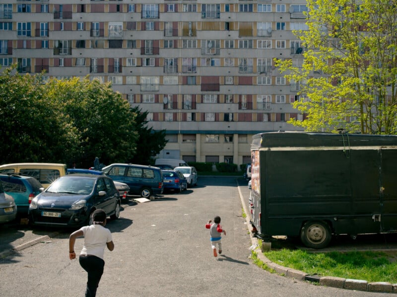 A child and adult run down a parking lot towards a large, colorful apartment building, with parked cars lining the sides and greenery in the background.