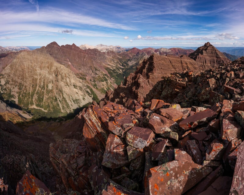 Rocky, reddish mountain peaks under a blue sky with scattered clouds; rugged foreground of layered rocks, with valleys and distant mountains visible in the background.