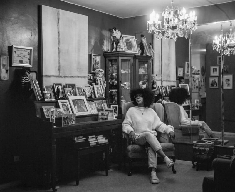 A person with an afro sits in an armchair in a room filled with family photos, a piano, and chandeliers. The walls have framed art and mirrors, reflecting the scene and adding depth to the cozy, nostalgic space.