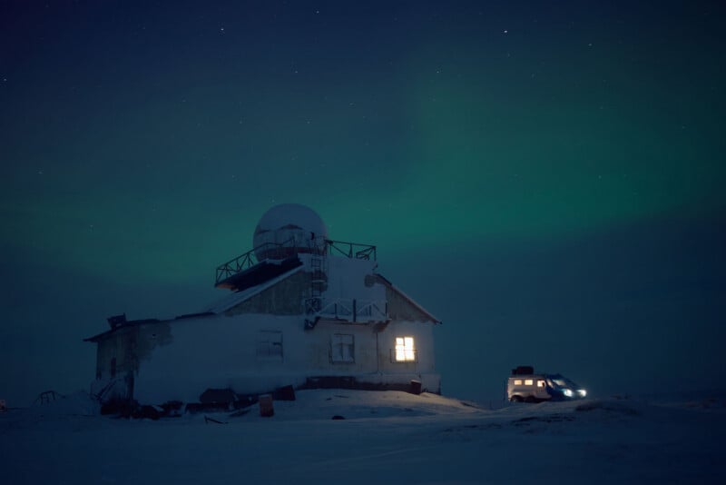 An old snow-covered building with lit windows stands in the dark, snowy landscape. A vehicle with headlights on is parked nearby. The sky is dark with a green aurora visible.