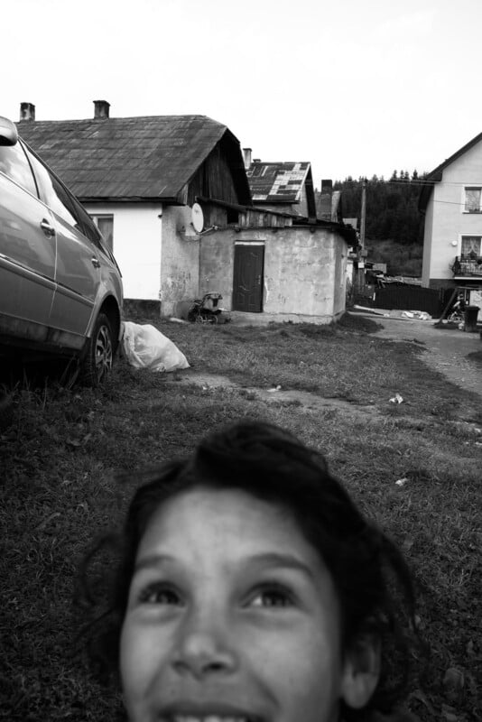 A smiling child in the foreground with houses, a car, and grassy yard in the background, shown in black and white.