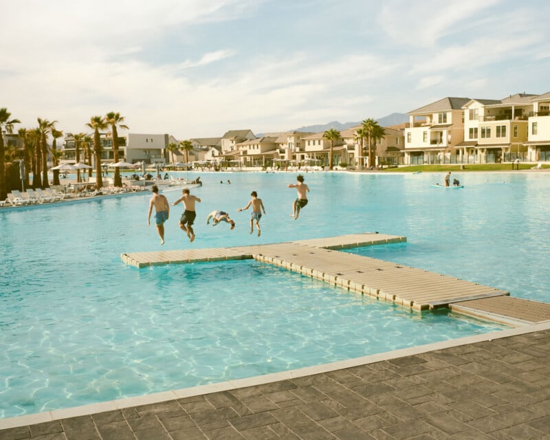 Five people jump off a wooden dock into a large, clear swimming pool, surrounded by modern houses and palm trees under a blue sky. Other people relax and swim in the background.