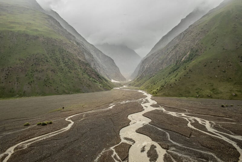 A braided river flows through a wide, rocky valley surrounded by steep, green mountains under a cloudy, misty sky.