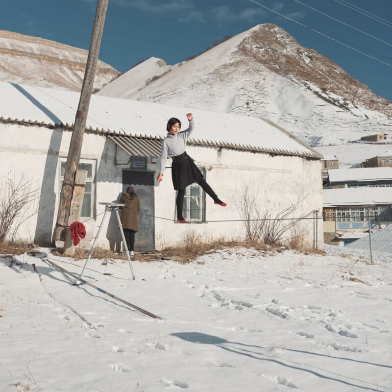 A person balances on a slackline in front of a snow-covered house with mountains in the background, while another person stands near the door.