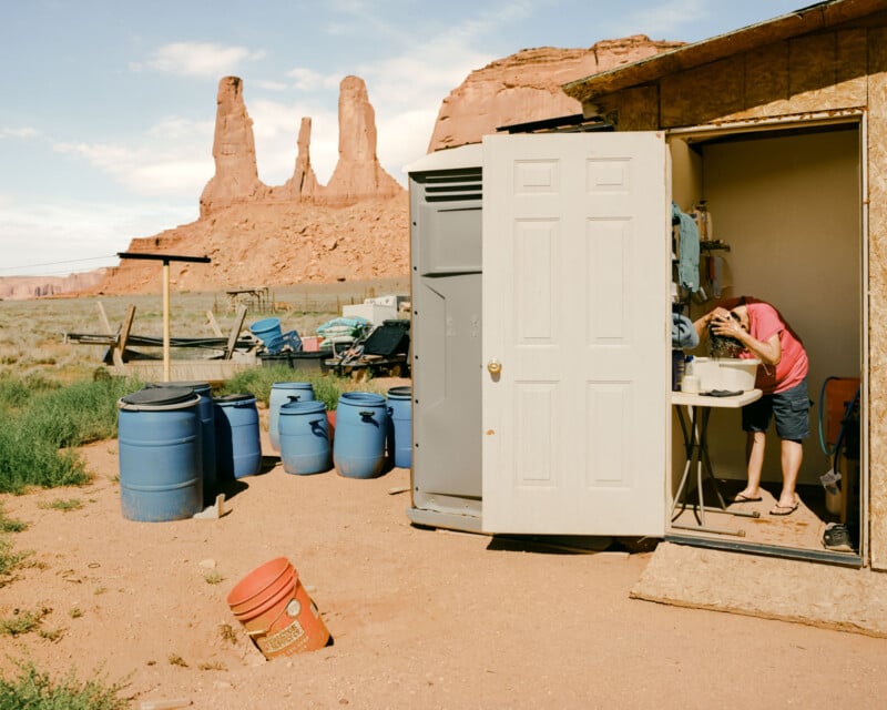 A person bends over a sink inside a small shed surrounded by blue barrels in a desert landscape with tall, twin rock formations in the background.