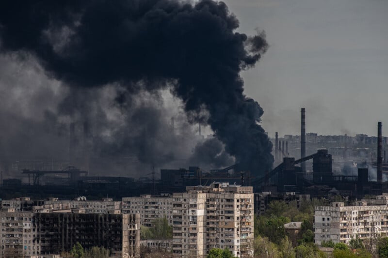 Thick black smoke rises from industrial buildings behind apartment blocks, indicating a large fire or explosion in an urban-industrial area. The sky is overcast, and the scene appears tense and hazardous.