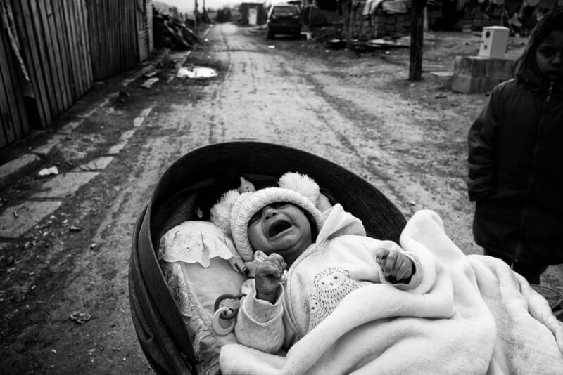 A crying baby lies bundled in a stroller on a rough, muddy street, with rundown structures and a child standing nearby. The scene is in black and white, evoking a somber, gritty atmosphere.