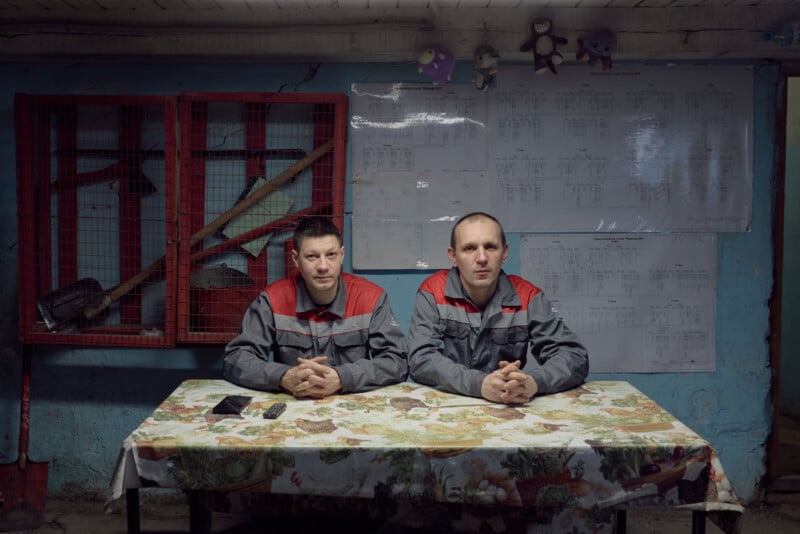 Two men in gray and red uniforms sit at a table with a floral tablecloth in a dimly lit room. Tools hang behind them, and papers are posted on a blue wall. Plush toys are lined up on a shelf above.
